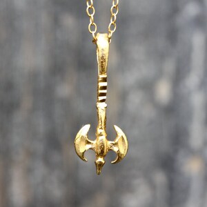 May include: A gold-colored necklace with a double-headed axe pendant. The axe has a detailed design and is suspended from a delicate chain.