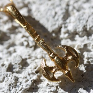 May include: A gold-colored necklace with a pendant shaped like a double-headed axe. The pendant has a textured surface and a detailed design.