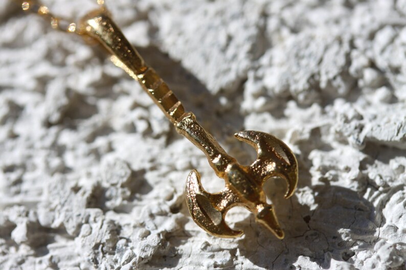 May include: A gold-colored necklace with a pendant shaped like a double-headed axe. The pendant has a textured surface and a detailed design.