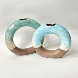May include: Two circular ceramic vases with a hollow centre. The larger vase features a teal and cream glaze over a brown base. The smaller vase has a light blue and cream glaze over a brown base. Both vases have a small opening at the top.