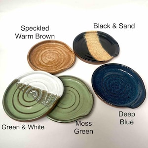 May include: A selection of handmade ceramic spoon rests in various colours. The rests include speckled warm brown, black and sand, green and white, moss green, and deep blue. Each rest has a small handle and a circular design.