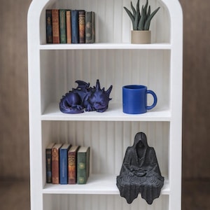 Puede incluir: Una estantería blanca con forma de arco que exhibe varios objetos decorativos. Incluye libros en miniatura, una taza azul, una figura de dragón púrpura durmiente, una figura negra con capucha y una pequeña planta en maceta.