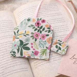 Op de afbeelding: Handgemaakte bloemen theezakje bladwijzer. De bladwijzer heeft een bloemenpatroon met roze, gele en groene bloemen op een witte achtergrond. Een roze lint is bevestigd. De bladwijzer ligt op een open boek.