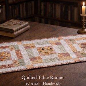 May include: A handmade quilted table runner, measuring 38 cm by 107 cm, with a patchwork design in cream, green, and rust. The runner is displayed on a dark wooden table, with a vase of flowers, books, and a lit candle.