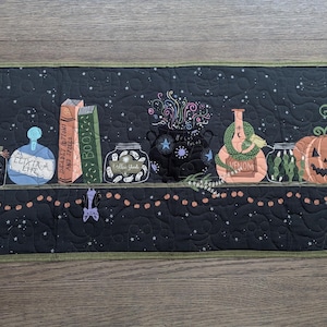 Puede incluir: Un camino de mesa rectangular acolchado en negro con motivos de Halloween bordados, incluyendo frascos de pociones etiquetados Elixir of Life, Deadly Potions and Spells y Venom, un caldero negro, una serpiente verde, una calabaza y un murciélago.