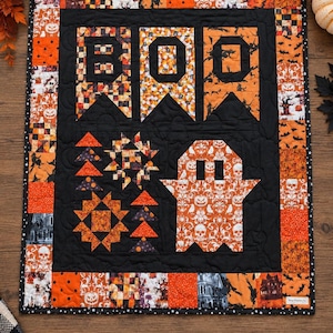 Puede incluir: Un edredón de pared rectangular con temática de Halloween que presenta un fondo negro con la palabra BOO en letras grandes. Incluye bordes de patchwork con patrones naranjas y negros, una forma de fantasma y motivos geométricos.