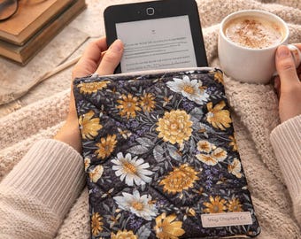 Op de afbeelding: Een e-reader in een bloemenpatroon hoes met gele en witte bloemen, vastgehouden door een persoon. De hoes ligt op een beige deken, met een kopje koffie en een kaars op de achtergrond. De tekst luidt "Perfect for every book lover."