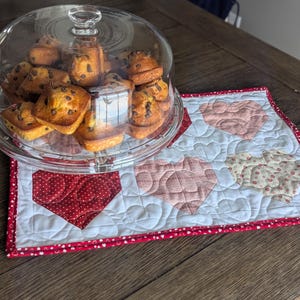 May include: A glass cake stand with a clear dome lid holds several chocolate chip muffins. The stand sits on a quilted placemat with heart-shaped designs in red, pink, and cream colors, on a light blue background.