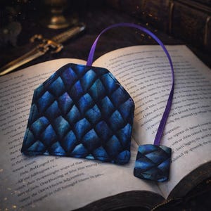 May include: A dragon scale-patterned bookmark with a matching tassel, in shades of blue and purple, rests on an open book. A candle and a decorative knife are in the background. The text at the bottom says, "For Readers Who Love Dragons & Dark Chapters."