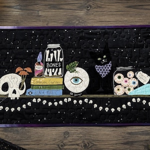 Puede incluir: Un tapiz rectangular de tela negra acolchada que muestra un estante con tubos de ensayo, una calavera con setas, un frasco etiquetado como BONES, libros titulados Common Curses, Spooky Spells y Black Magic, una rana sobre un globo ocular y un gato.