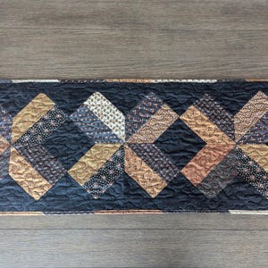 May include: A rectangular quilted table runner featuring a repeating geometric pattern. The design incorporates various fabrics in shades of brown, gold, and navy blue, set against a black background. The runner measures approximately 36 inches long.