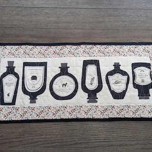 Puede incluir: Un camino de mesa acolchado rectangular con un panel central color crema que presenta seis ilustraciones de frascos de boticario negros, cada uno con etiquetas y texto únicos, rodeado por un borde floral con pequeñas flores rojas y azules.