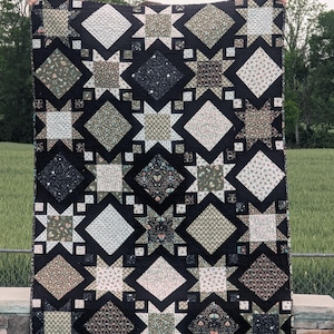 May include: A handmade quilt with a black background and a repeating star pattern. The stars are filled with various floral and geometric patterns in shades of green, cream, and black. The quilt is displayed outdoors.