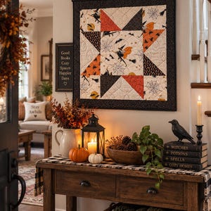 May include: A square quilted wall hanging featuring a star pattern with orange, black, and cream fabric printed with witches, bats, and spiderwebs. The quilt is framed by a black border and hangs from a metal rod on a wall above a wooden console table.