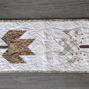 May include: A rectangular quilted table runner featuring a white background with a stitched leaf pattern. Four fabric leaf appliques in shades of gold, brown, plaid, and orange are centered horizontally, each with a dark brown fabric stem.