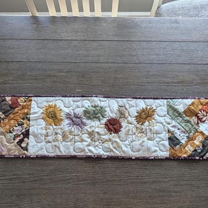 May include: A rectangular quilted table runner featuring a patchwork design with autumn-inspired colours including orange, brown, yellow, and cream. The centre panel displays embroidered sunflowers and the word "thankful" in cursive script.