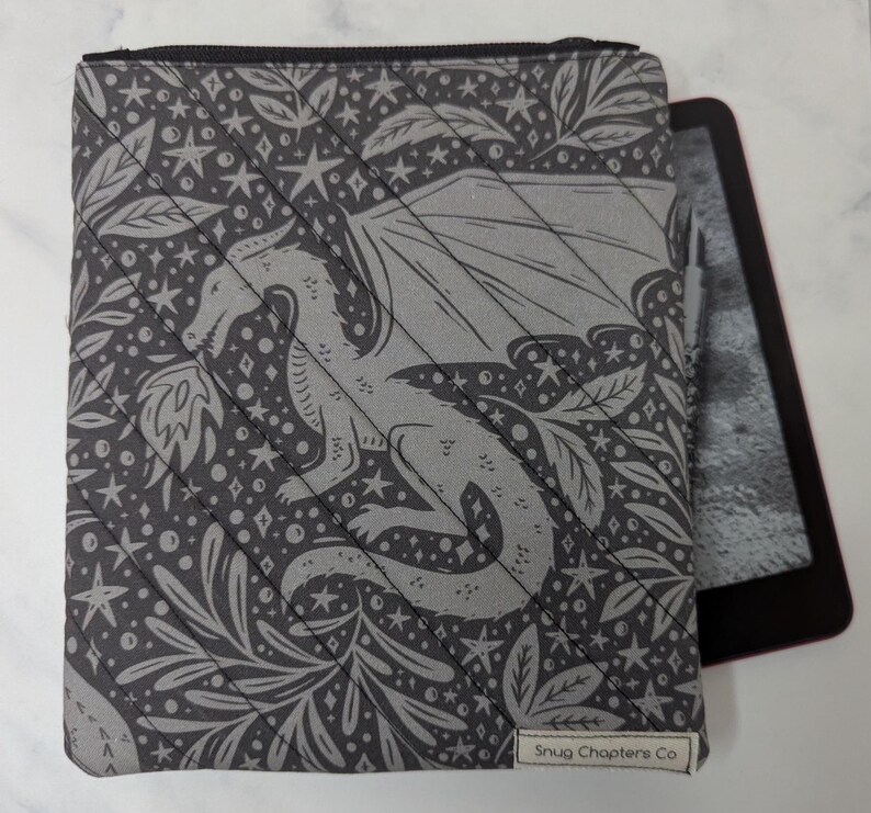 Op de afbeelding: Een gewatteerde grijze stoffen hoes met een donkergrijs patroon van een draak, sterren en bladeren. De hoes heeft een zwarte rits aan de bovenkant en een klein wit label rechtsonder met de tekst Snug Chapters Co. Een e-reader is zichtbaar.