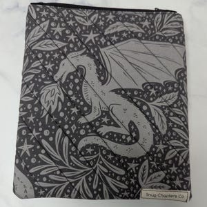 Op de afbeelding: Een rechthoekig stoffen tasje met een zwarte ritssluiting en een diagonaal doorgestikt patroon. De grijze stof is bedrukt met een donkergrijs motief van een draak, sterren en bladeren. Een klein wit label bevat de tekst Snug Chapters Co.
