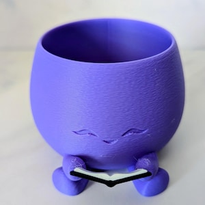 Op de afbeelding: Een paarse 3D-geprinte plantenbak in de vorm van een personage met een lachend gezicht en een boek. De plantenbak heeft een afgerond lichaam, twee kleine voetjes en een brede opening aan de bovenkant voor planten. Het boek is zwart en wit.