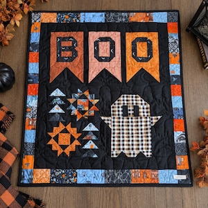 Könnte beinhalten: Ein rechteckiger Wandbehang aus Stoff mit schwarzem Hintergrund und dem Wort BOO in orangefarbenen und schwarzen Buchstaben. Das Design umfasst ein schwarz-weiß kariertes Gespenst, orangefarbene Sternmuster und einen Rand aus bunten Quadraten.