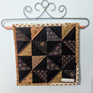 May include: A small, square quilt with a patchwork design of black and patterned triangles. The quilt is framed with a floral patterned border and is hanging on a decorative metal rack. The quilt measures approximately 10 inches square.