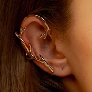 May include: A gold-colored ear cuff featuring a branch-like design. The ear cuff curves around the ear, with multiple antler-like projections. The jewelry is worn on the ear and is set against a background of brown hair and skin.