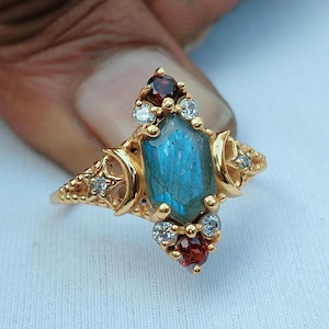 May include: A gold ring featuring a central hexagonal blue labradorite stone, flanked by two crescent moon motifs. The setting includes small round red garnet stones at the top and bottom, accented by tiny clear white stones along the band.