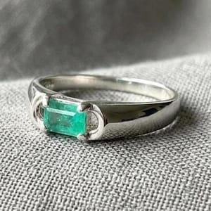 Könnte beinhalten: Ein silberfarbener Ring mit einem einzelnen, im Smaragdschliff geschliffenen grünen Edelstein, der horizontal in der Mitte gefasst ist. Die Ringschiene ist glatt und poliert und besitzt eine geschwungene Fassung, die den Stein einrahmt.