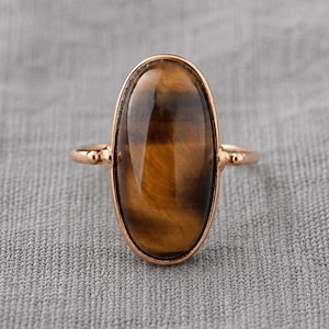Op de afbeelding: Een ring met een grote, ovaalvormige tijgeroog-edelsteen met bruine en gouden chatoyante banden. De steen is gezet in een gepolijste roségoudkleurige metalen band met een eenvoudig, slank ontwerp en kleine decoratieve accenten aan de zijkanten.