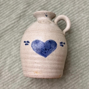 May include: A small, off-white ceramic jug with a blue heart painted on the front. The jug has a handle and a narrow spout. Decorative blue dots flank the heart. The jug has a rustic, handmade appearance.