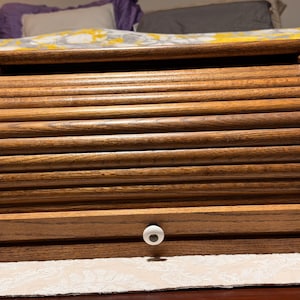 May include: A wooden bread bin with a roll-top lid and a drawer. The bread bin is made of light brown wood with a natural grain. The drawer has a white knob. The bread bin is designed to store bread and baked goods.