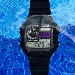 May include: A black Casio digital watch with a black resin strap, featuring a square face with a digital time display, a world map graphic, and a circular inset showing a black cat. The watch face displays the text WR100M and 10 YEAR BATTERY.