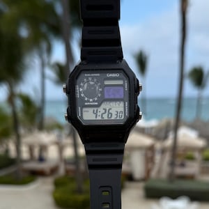 May include: A black Casio digital watch with a rectangular face, featuring a black resin strap, four side buttons, and a digital display showing the time 4:26:30, the text NYC, and a small circular graphic with a cat silhouette in the upper left corner.