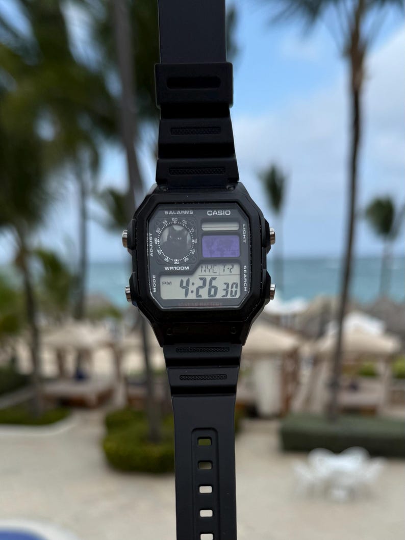 May include: A black Casio digital watch with a rectangular face, featuring a black resin strap, four side buttons, and a digital display showing the time 4:26:30, the text NYC, and a small circular graphic with a cat silhouette in the upper left corner.
