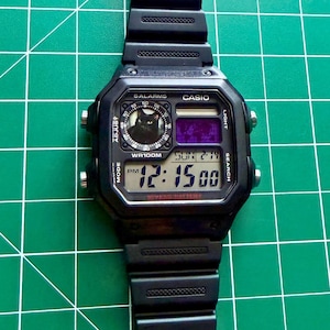 May include: A black Casio digital watch with a resin strap and a square case. The watch face features a small circular graphic of a black cat, a purple digital display, and text including 5 ALARMS, WR100M, and 12:15 PM on the main screen.
