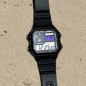 May include: A black Casio digital watch with a square face, featuring a world map display, multiple alarm settings, and a 10-year battery indicator. The watch has a black resin strap and four silver-tone side buttons for adjusting settings.