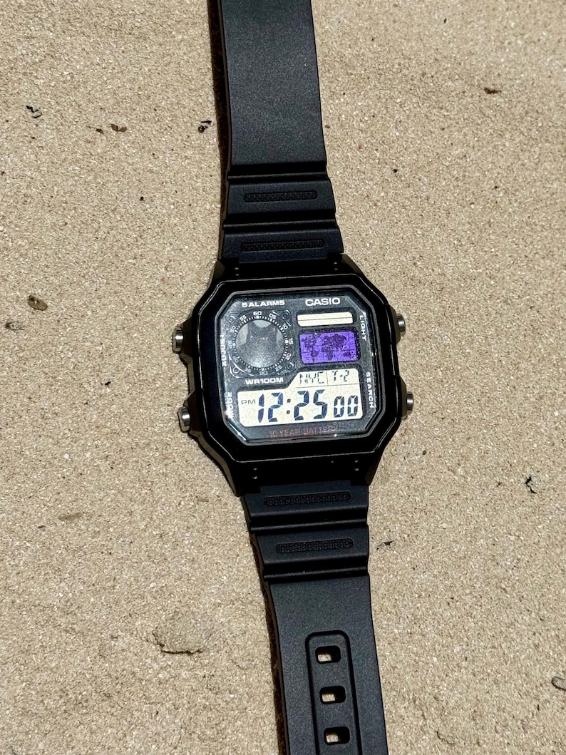 May include: A black Casio digital watch with a square face, featuring a world map display, multiple alarm settings, and a 10-year battery indicator. The watch has a black resin strap and four silver-tone side buttons for adjusting settings.