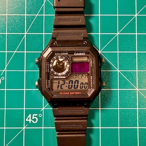 May include: A black Casio digital watch with a resin strap, featuring a square face with a black cat graphic in the circular sub-dial, a digital time display showing 12:00, the text 10 YEAR BATTERY, and four silver-tone side buttons for control.