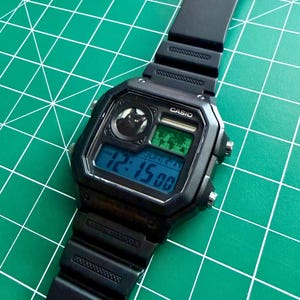 May include: A black Casio digital wristwatch with a rectangular case and a black resin strap. The watch face features a small circular graphic of a black cat, a world map display, and a digital screen showing the time 12:15 and the date SUN 2-14.