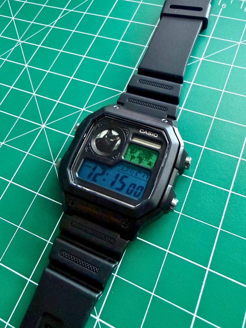 May include: A black Casio digital wristwatch with a rectangular case and a black resin strap. The watch face features a small circular graphic of a black cat, a world map display, and a digital screen showing the time 12:15 and the date SUN 2-14.