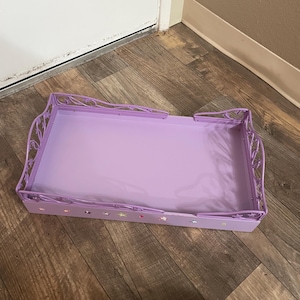May include: A rectangular, lavender metal tray with decorative filigree edges. The tray has a solid lavender base and is decorated with small, colorful star-shaped embellishments along the sides. The tray is designed for decorative or functional use.