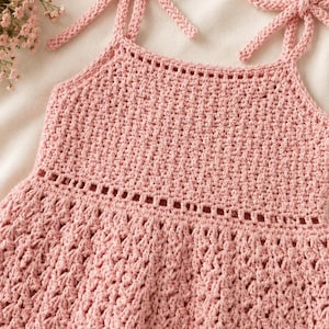 Pode incluir: Um vestido rosa claro, feito à mão em crochê, com alças amarradas. O vestido apresenta um padrão de ponto detalhado e uma saia rodada. As alças são amarradas em laços nos ombros. O vestido é feito de fio macio.