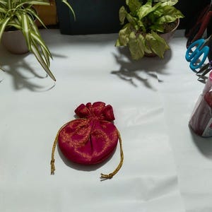 Può includere: Una piccola borsa rotonda color magenta con coulisse e ricami dorati, posizionata al centro. A sinistra ci sono diverse caramelle incartate in oro. A destra, tre rocchetti cilindrici di filo rosso, verde e blu, insieme a delle forbici.