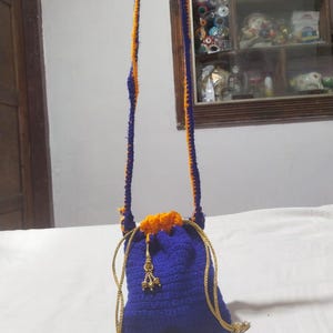 Pode incluir: Uma bolsa tipo saco de crochê em azul royal com uma alça longa a condizer. A bolsa apresenta uma borda superior de crochê em laranja brilhante e cordões trançados dourados. Um pequeno pendente de metal dourado com três sinos pendurados.