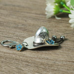 May include: Silver-tone metal keychain featuring a miniature sombrero design. The hat has a decorative pattern and is accented with blue floral details. Includes a lobster clasp for easy attachment.