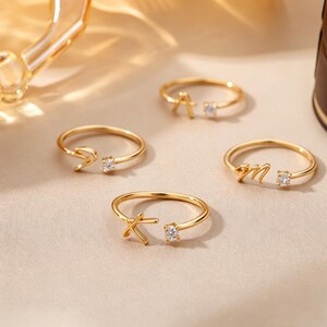 May include: Four gold-toned open rings, each featuring a single letter and a small clear stone at the ends. The letters displayed are A, S, M, and K, rendered in a cursive or script font. The rings have a thin, polished metal band design.
