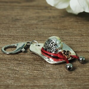 May include: A silver-toned keychain with a decorative charm. The charm features a small, ornate hat with a red cord, a small gold-colored charm, and two silver-colored beads. The keychain has a silver clasp.