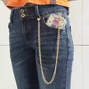 May include: A gold-colored chain attached to a clear phone case with a cartoon design, hanging from the pocket of blue denim jeans. The jeans have a double-button closure and are worn with an orange top.