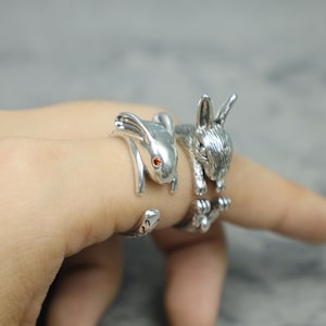 May include: A silver-toned open ring featuring two rabbit figures facing opposite directions. One rabbit has a smooth finish with a small orange stone set as an eye, while the other rabbit has textured fur detailing and visible paws at the bottom.