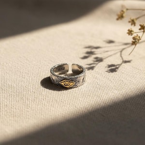Può includere: Un anello in argento regolabile caratterizzato da un motivo centrale a occhio color oro circondato da raggi solari incisi e motivi geometrici. La fascia interna è chiaramente stampata con il testo 5925, che indica la composizione del metallo.